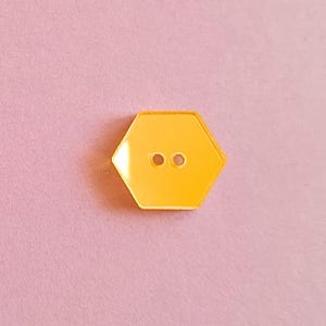 Puede incluir: Un botón hexagonal amarillo brillante con dos pequeños agujeros. El botón está centrado en un fondo rosa suave. Este botón es un suministro de costura o artesanía.