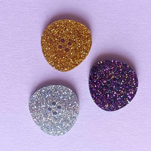 Peut inclure: Trois boutons ovales scintillants en or, argent et violet. Chaque bouton comporte quatre petits trous pour la couture. Les boutons sont posés sur un fond violet clair.