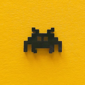 Puede incluir: Nave espacial alienígena pixelada negra con un diseño simple. La nave espacial está sobre un fondo amarillo.