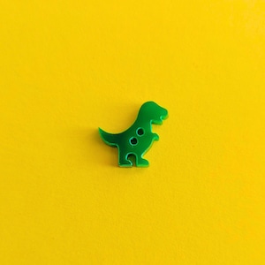 May include: A bright green dinosaur-shaped button with two small holes, set against a vibrant yellow background. The button is a simple, playful design, perfect for sewing or craft projects.