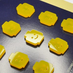 May include: Nine yellow plastic buttons shaped like Lego minifigures with smiling faces. The buttons are designed and handmade in France.