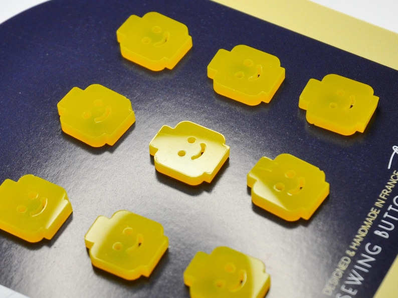 May include: Nine yellow plastic buttons shaped like Lego minifigures with smiling faces. The buttons are designed and handmade in France.