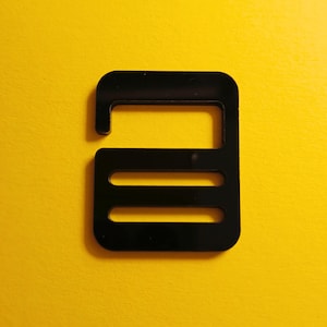 Peut inclure: Boucle en plastique noire de forme rectangulaire avec trois fentes horizontales. La boucle a une petite boucle en haut.