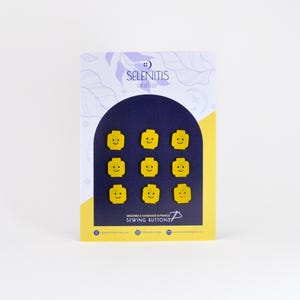 May include: A set of nine yellow sewing buttons shaped like Lego minifigure heads with smiling faces. The buttons are displayed on a card with the text "Selenitis atelier Sewing Buttons".