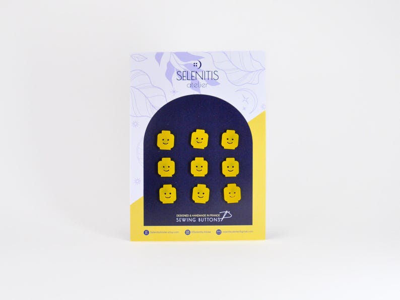 May include: A set of nine yellow sewing buttons shaped like Lego minifigure heads with smiling faces. The buttons are displayed on a card with the text "Selenitis atelier Sewing Buttons".