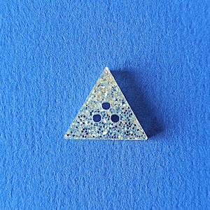 Puede incluir: Un botón de resina transparente de forma triangular con un acabado de purpurina plateada y tres puntos de purpurina azul en el centro.