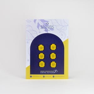 May include: A set of six yellow sewing buttons shaped like smiling Lego minifigure heads. The buttons are displayed on a card with the text "SELENITIS atelier" and "SEWING BUTTONS".