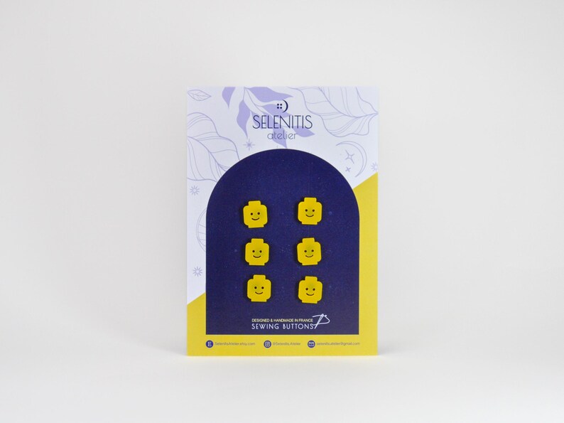 May include: A set of six yellow sewing buttons shaped like smiling Lego minifigure heads. The buttons are displayed on a card with the text "SELENITIS atelier" and "SEWING BUTTONS".