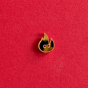 Puede incluir: Un pin de esmalte dorado con forma de llama, con un contorno negro y dos pequeños círculos rojos. El pin está sobre un fondo rojo.