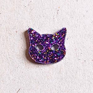 May include: A purple glitter cat-shaped embellishment with two eye holes. The cat has pointed ears and is covered in iridescent glitter. The background is a light, textured surface.