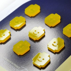May include: Eight yellow plastic buttons shaped like Lego minifigures with smiling faces. The buttons are arranged in a circle on a blue background.