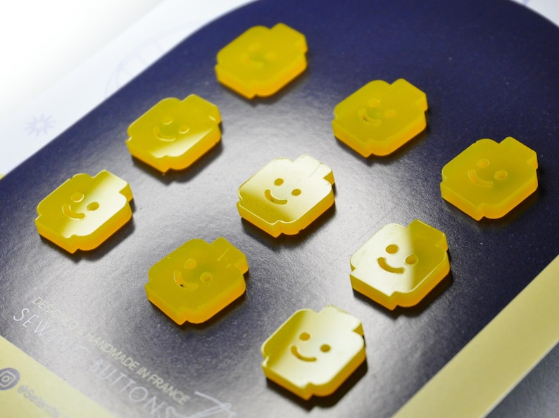 May include: Eight yellow plastic buttons shaped like Lego minifigures with smiling faces. The buttons are arranged in a circle on a blue background.