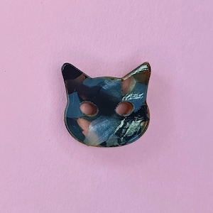 May include: A black cat face shaped pin with a blue, green and pink abstract pattern.