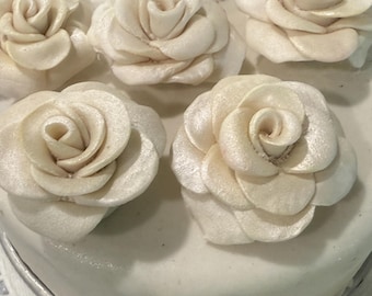 May include: A close-up of a white cake decorated with five white sugar roses. The roses are arranged in a circular pattern around the cake.