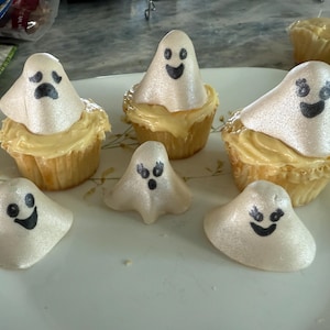 Puede incluir: Cupcakes con temática de Halloween en un plato. Los cupcakes tienen glaseado amarillo y están cubiertos con merengues blancos en forma de fantasma con caras negras. Los fantasmas tienen diferentes expresiones.