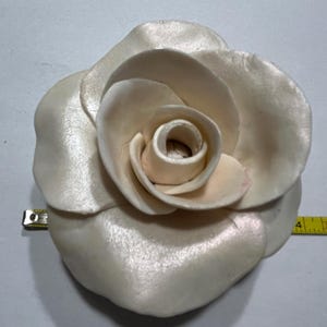 Könnte beinhalten: Eine große, weiße Satinrose mit perlmuttartigem Schimmer. Die Rose ist aus Stoff gefertigt und hat ein realistisches Aussehen.