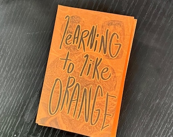 May include: A stack of orange-colored cards with the text "Learning to like ORANGE" in black, handwritten-style lettering. The background features a faint orange slice design.