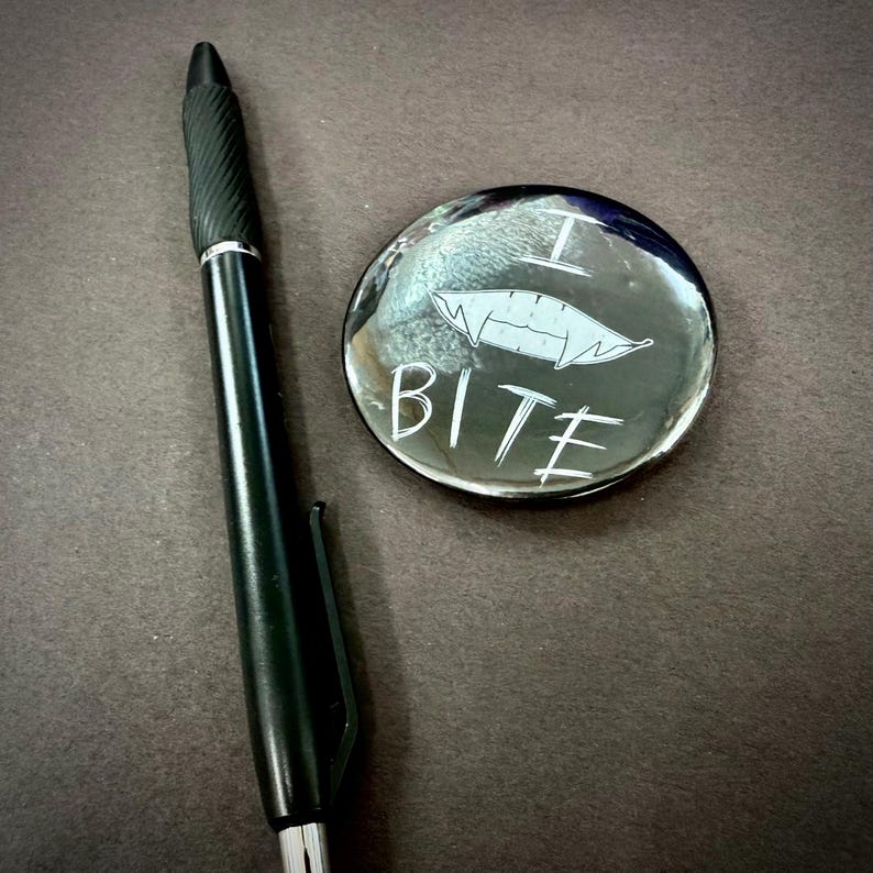 May include: A black pen with a textured grip rests beside a round, glossy button. The button features a stylized set of fangs and the word "BITE" in white lettering, creating a playful, gothic aesthetic.