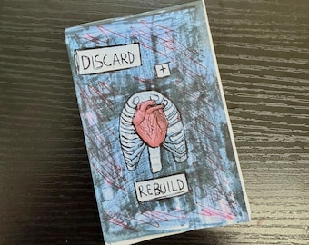 Puede incluir: Un pequeño libro azul y blanco con un corazón rojo y las palabras "DISCARD + REBUILD" en la portada.
