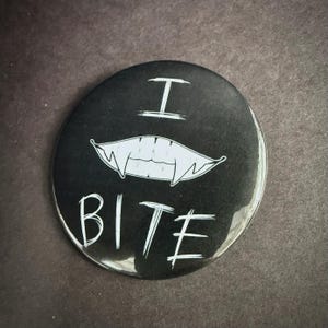 May include: Black button with white text and a vampire teeth graphic. The text reads "I BITE" in a handwritten style. The teeth are white and stylized. The button is round and has a glossy finish.