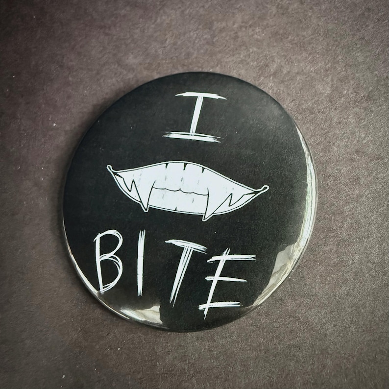 May include: Black button with white text and a vampire teeth graphic. The text reads "I BITE" in a handwritten style. The teeth are white and stylized. The button is round and has a glossy finish.