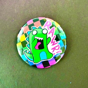 May include: A round button with a cartoon dinosaur illustration. The green dinosaur has a wide open mouth and the word "RAWWR!" in pink. The background is a colorful checkered pattern of pink, green, blue, and black.