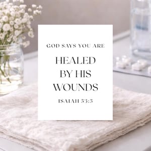 Peut inclure: Une carte blanche avec le texte "GOD SAYS YOU ARE HEALED BY HIS WOUNDS ISAIAH 53:5" repose sur un tissu beige plié. La carte est centrée, avec le texte dans une police simple et élégante. Un bocal en verre avec des fleurs blanches est visible en arrière-plan.