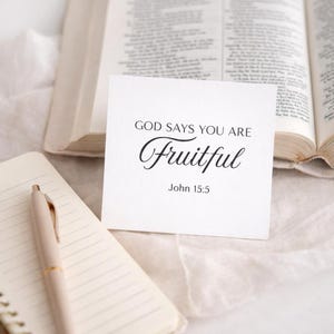 May include: A white card with the text "GOD SAYS YOU ARE Fruitful" and "John 15:5" in black script. The card is placed in front of an open book and a notepad with a pen.