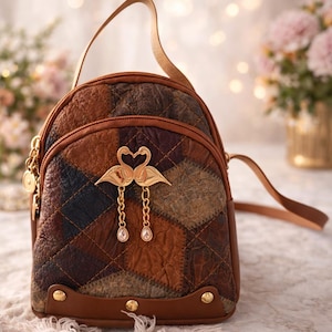 Può includere: Uno zaino patchwork marrone con un'emblema di cigno dorato. Il piccolo zaino ha un manico superiore e spallacci regolabili. La borsa è realizzata in pelle e ha finiture dorate. La borsa misura circa 25 cm di altezza.