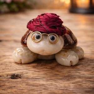 Può includere: Una stravagante statuetta a forma di tartaruga con testa bianca, corpo beige e guscio marrone. Indossa un cappello bordeaux e occhiali con montatura dorata. La statuetta è esposta su una superficie di legno, creando un oggetto decorativo unico.