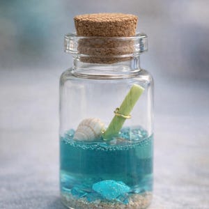 Puede incluir: Una botella de vidrio transparente con tapón de corcho, llena de líquido azul, arena y pequeños cristales azules. En el interior, una concha y un pergamino sujeto por un anillo dorado. Tema náutico.