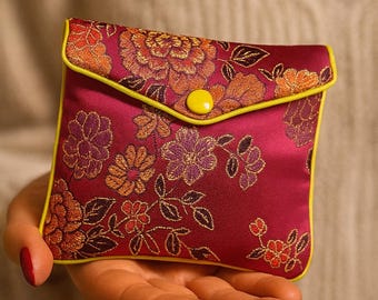 May include: A small, square pouch with a floral pattern in shades of burgundy, orange, and purple. The pouch has a yellow trim and a yellow button closure. The design is reminiscent of traditional Asian textiles.