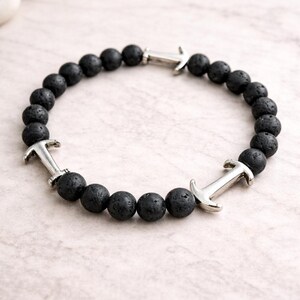 Könnte beinhalten: Ein Armband aus schwarzen Lavaperlen mit silberfarbenen Ankeranhängern. Das Armband hat runde, strukturierte schwarze Perlen und drei Ankeranhänger, die gleichmäßig über den Umfang verteilt sind. Das Armband wird auf einer hellrosa und weißen Oberfläche präsentiert.