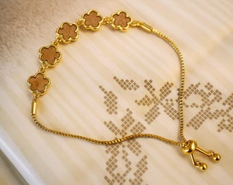 May include: A gold-colored bracelet featuring flower-shaped charms with a light brown center. The bracelet has a delicate chain and an adjustable clasp. The jewelry is displayed on a light-colored surface with a subtle pattern.