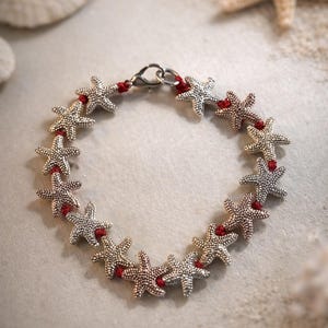 Op de afbeelding: Een zilveren armband met zeesterren en een rood koord. De armband bestaat uit meerdere zeesterbedels die zijn verbonden door een rood koord en een zilveren sluiting. De achtergrond toont schelpen en zand.