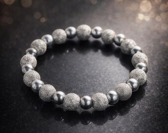 Puede incluir: Una pulsera plateada con una mezcla de cuentas texturizadas y lisas. La pulsera está hecha de pequeñas cuentas redondas en varios tonos de plata y gris. La pulsera se muestra sobre una superficie oscura y reflectante.