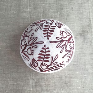 May include: White kippah with a burgundy floral embroidery design. The embroidered pattern features stylized leaves and flowers, creating a decorative and detailed appearance. The kippah is round and sits on a neutral-colored fabric.