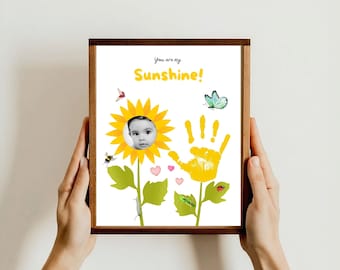 Może przedstawiać: Wydruk w ramie z żółtym odciskiem dłoni w kształcie słonecznika. Słonecznik ma twarz dziecka w środku i napis "You are my Sunshine!" u góry. Wydruk jest ozdobiony małymi obrazkami motyli, pszczół, biedronek i gąsienic.