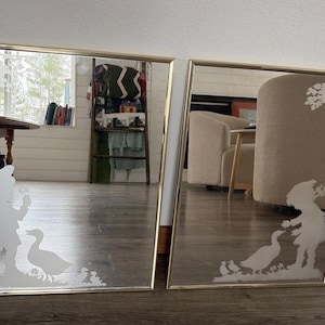 Op de afbeelding: Twee rechthoekige spiegels met gouden frames. Elke spiegel heeft een mat ontwerp van een kind met een mand, een eend en eendenkuikens. De spiegels weerspiegelen het interieur van de kamer, inclusief meubels en decoratie.