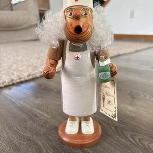 Puede incluir: Figurita de madera de una enfermera con un vestido blanco, un gorro con una cruz roja y gafas. Lleva una botella verde con la etiqueta "Ol. Ricino" y una etiqueta blanca con texto en alemán.