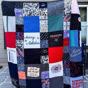 May include: A memory quilt constructed from various fabric squares featuring diverse patterns, colors, and printed text including Kenny Robertson, Grandpa Rocks, Out of My Mind Back in 5 Minutes, and Rummel Seniors Class of 69, with black stitched borders.