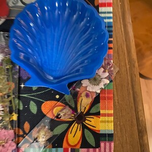 May include: A blue plastic seashell-shaped dish on a colorful fabric background.
