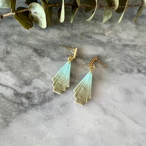 May include: A pair of Art Deco-style earrings. Each earring features a fan-shaped design with a gradient of light blue to beige. The earrings have gold-colored posts and accents, set against a marble background.