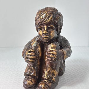 May include: A bronze sculpture of a young person sitting with their legs crossed. The sculpture has a distressed finish, giving it an aged look.
