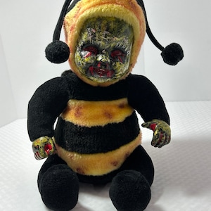May include: A plush toy shaped like a bee with a creepy, zombie-like face. The bee is wearing a yellow and black striped costume with a black and yellow striped hat. The toy has red painted details on its hands and face.