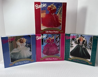 May include: Three boxed 120-piece Barbie limited edition Christmas puzzles. Each box features a different Barbie doll in a festive gown, with a snowy winter scene in the background. The boxes are red, pink, and green.