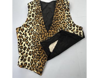 Op de afbeelding: Een mouwloos vest met een bruin en zwart luipaardprintpatroon op de voorpanden. Het kledingstuk heeft een diepe V-hals, een enkele zichtbare knoopsluiting en een effen zwarte binnenvoering zichtbaar op de omgevouwen voorflap.