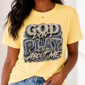 Puede incluir: Una camiseta de manga corta de color amarillo pálido con un estampado gráfico en el pecho. El diseño muestra el texto "GOD DON'T PLAY ABOUT ME" en una fuente llamativa, metálica y brillante con un fondo de textura de mezclilla azul.