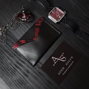 May include: A black leather cardholder with a red snakeskin accent, a silver watch with a red face, and a silver pendant with a red gem are displayed on a dark wooden surface. A business card with the text "ATER SALUS Handmade Leathergoods" is also present.