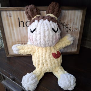 May include: A handmade, plush bunny doll with a yellow body, white head and feet, and brown ears. The doll has a red heart on its chest and closed black eyes. The background includes a wooden sign with the word "home".